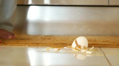 Serin mutfak zemininde çıplak ayakla duran küçük bir çocuk, kırık bir yumurtanın yanında merakla bakıyor.