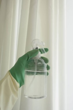 A person is holding a spray bottle intended for cleaning, positioned near beautiful white curtains clipart