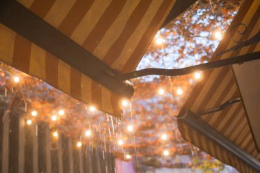 Turuncu bir gölgelik hafif yağmurla örtülmüş, fenerler ve yumuşak ışıklar sıcaklık yaratıyor.