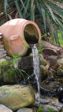 Güzel işlenmiş büyüleyici bir kil tenceresi sakin bir bahçeye nazikçe su döküyor.