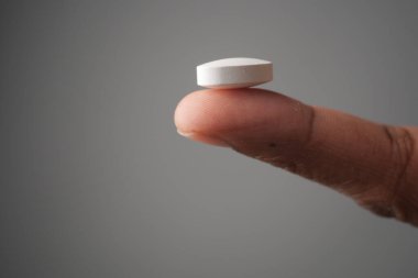 Küçük, beyaz bir hap, parmak ucuyla yumuşak gri bir arka plan arasında hassas bir şekilde dengelenir.