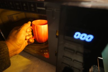 Bir el, mikrodalga fırından sıcak bir içecek dolu neşeli kırmızı bir kupayı nazikçe çeker.