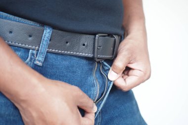 Siyah kuşak ve siyah gömlek giyerken bir insan mavi kotunun fermuarını ayarlar. Bu hareket, muhtemelen evde olan sıradan bir ev ortamını işaret ediyor..