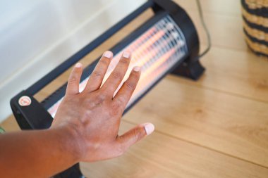 Bir el, ahşap bir zemindeki ısıtıcıya doğru uzanıyor. Isıtıcı yumuşak ışık yayarak serin havanın ortasında sıcak bir atmosfer yaratıyor..