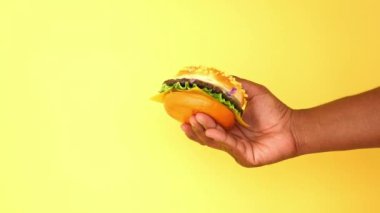 Bir insan elinde renkli ve yaratıcı bir hamburgerle cesur sarı bir zemin sunar..