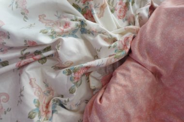 Açık renkli ve pembe aksanlı yumuşak çiçekli bir yatağın yakın görüntüsü. Kumaş buruşmuş görünüyor, ortama rahatlatıcı ve davetkar bir hava katıyor..