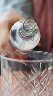 Bir el kademeli olarak cam bir şişeyi eğerek şeffaf bir sıvının güzelce işlenmiş kristal bir bardağa akmasını sağlar. Ortalık sakin ve davetkar..