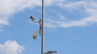 Uzun bir direğe monte edilmiş iki güvenlik kamerası bulutlarla dolu açık mavi bir gökyüzünün altındaki hareketli bir şehir bölgesindeki aktiviteyi kaydediyor. Gözetleme teknolojisi halkın güvenliğini arttırıyor.