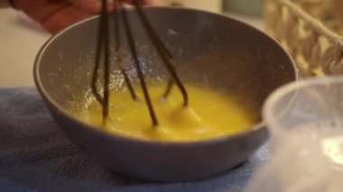 Bir kişi bir kasede yumurta çırparak yemek hazırlıyor. Mutfak sıcak ve davetkar, yakınlarda çeşitli yemek aletleri var. Süreç ayrıntılara dikkat ettiğini gösteriyor..