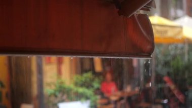 Sıcak bir kafe atmosferinde müşteriler şemsiyelerinin altında içkilerinin tadını çıkarırken, çatı güneşliğinden asfalta yağmur yağıyor. Canlı yeşillik ortama cazibe katıyor..