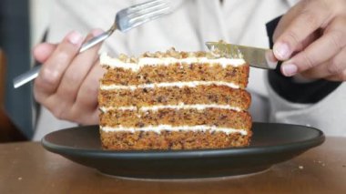 Bir insan havuçlu kekin tadını çıkarıyor, çatal ve bıçakla katmanları kesiyor. Tatlıda krema ve fındık var, koyu bir tabakta..