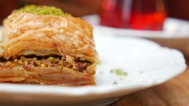 Baklava, yanında aromatik çay bulunan beyaz bir tabakta servis edilir. Ortam sıcak ve davetkar, tatlı bir ikram için mükemmel. Bu klasik tatlının zengin lezzetlerinin ve dokusunun tadını çıkarın..