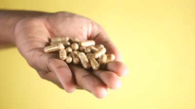Sarı bir arka planda çeşitli bitkisel kapsülleri gösteren bir elin yakın görüntüsü.