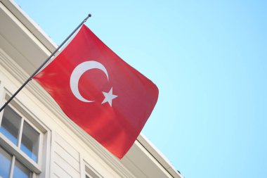 Açık gökyüzünün altındaki beyaz bir binanın önünden parlak bir Türk bayrağı geçiyor.