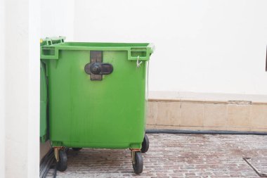 Şehir merkezinde bir duvarın yanında yeşil bir çöp konteyneri duruyor..