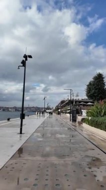 İnsanlar geniş bulutlu bir gökyüzünün altında rıhtımda geziniyor..