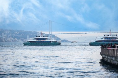 İki feribot suda ilerliyor ve arka planda bir köprü görünüyor..