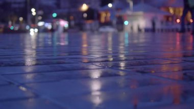 Şehir ışıkları akşamı aydınlatırken kaldırımda yağmur damlaları parlıyor..