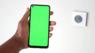 Bir kişi termostatın yanındaki yeşil ekranlı akıllı telefonu tutar..