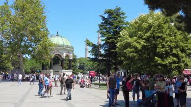 İnsanlar ağaçlarla çevrili halka açık bir meydanda güneşli bir günün tadını çıkarıyorlar..