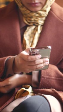 Kahverengi paltolu bir kadın ev ortamında telefonuna bakar..