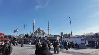 Kalabalık açık bir alanda, açık gökyüzünün altındaki sembolik bir caminin yanında hareket ediyor..