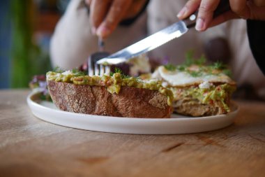 Bir insan avokadolarla kaplı açık yüzlü taze bir sandviç kesiyor..