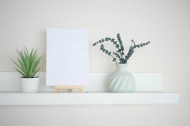 A simple shelf features a blank canvas, a plant, and a vase of greenery.