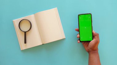 Bir insan açık bir kitap ve büyüteç yanında akıllı telefon tutar..