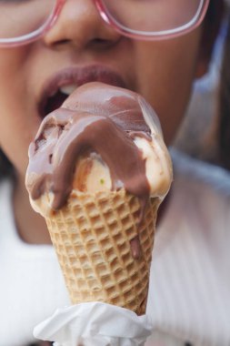 Küçük bir çocuk açık havada eriyen çikolatalı dondurma külahını ısırıyor..