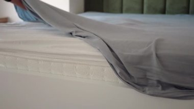 Bir insan açık gri bir çarşafı temiz, yapılmamış bir yatağa yerleştirir..