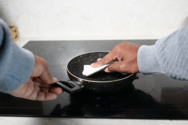 Bir insan yemek pişirirken tavayı indüksiyon tenceresine siler..