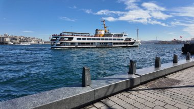 Bir feribot dalgalı sularda yol alır mavi gökyüzünün altında bir şehir zeminiyle.