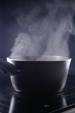 Düşük ışıkta yansıtıcı bir mutfak yüzeyine yerleştirilmiş bir çömlekten buhar yükselir..