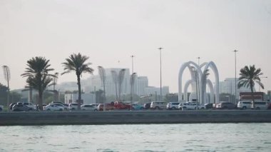 Araçlar, palmiye ağaçları olan bir rıhtım parkının yanındaki işlek bir yolda ilerliyorlar..