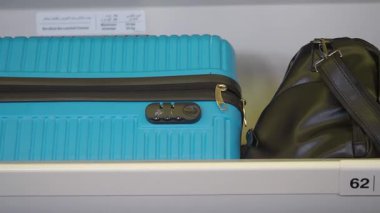 Parlak mavi bir bavul siyah bir çantanın yanındaki rafta sergileniyor..