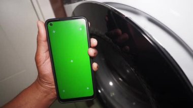 Birisi çamaşır makinesinin yanında yeşil ekranlı bir akıllı telefon gösteriyor..