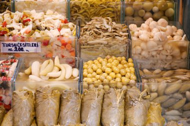 Sergide kalabalık bir pazarda kavanozlarda çeşitli sebzeler sergileniyor..