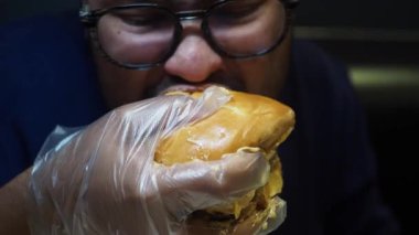 Bir adam, yiyeceksiz eldiven giyerken sulu bir burgeri ısırıyor..