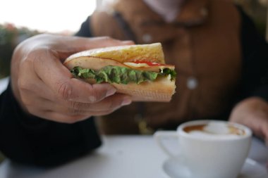 Bir insan kahve yudumlarken sebzelerle dolu taze bir sandviç tutar..