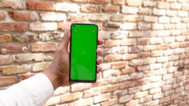 Kişi tuğlalara karşı boş yeşil ekranlı bir akıllı telefon gösterir.