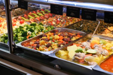 Bir açık büfe masasında renkli sebzeler ve sıcak yemekler sergilenir..