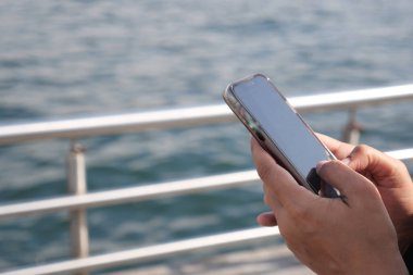 Bir kişi rıhtımda suyun yanında dururken elinde akıllı telefon tutuyor..