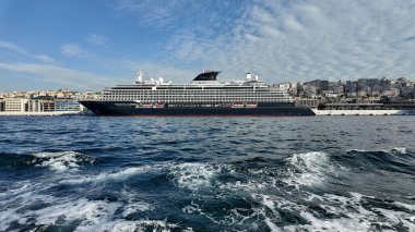 Büyük bir yolcu gemisi açık bir gökyüzünün altında hareketli bir limanda demirlemiş..