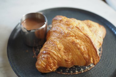 Altın kruvasan bir fincan sıcak çikolatanın yanında siyah bir tabağa konur..