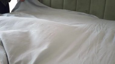 Bir insan zarif bir şekilde yatağın üzerindeki gri battaniyeyi düzleştiriyor..