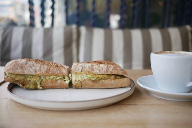 Yumurtayla dolu bir sandviçin iki yarısı bir fincan kahvenin yanında oturur..