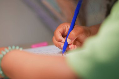 Bir çocuk masasında mavi bir kalemle yazmaya odaklanır, kendini işine verir..