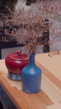 Mavi bir vazo, masadaki kırmızı bir konteynırın yanında kurumuş çiçekleri tutar..