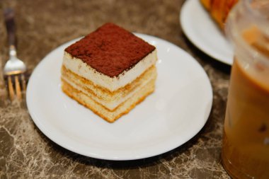 Zengin katmanlı bir tatlı beyaz bir tabakta bir içeceğin yanında oturur..